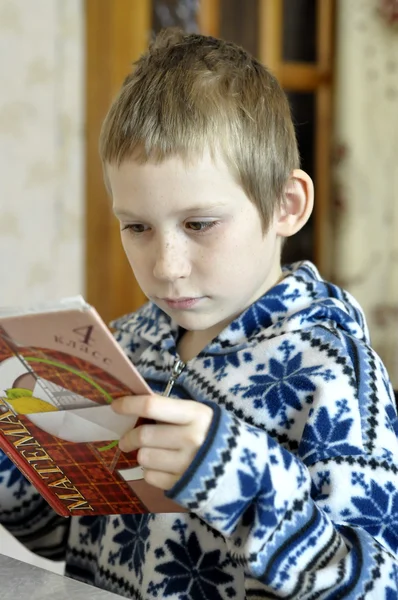 The 10-year-old boy sits with the textbook, doing homework. — Stock Photo, Image