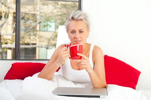 Portrait of a pretty woman sitting at her bed with a laptop and — Stock Photo, Image