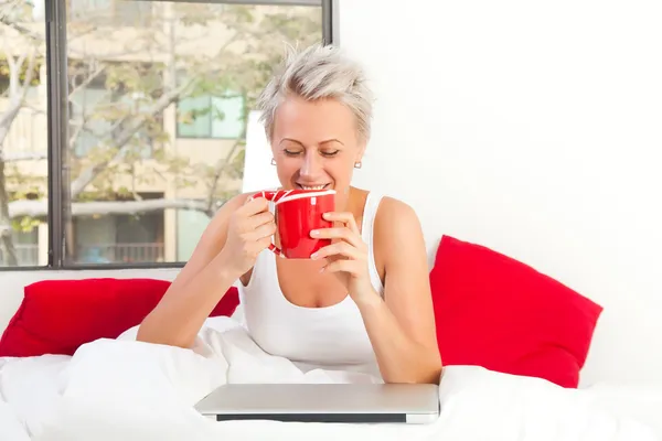 Portrait of a pretty woman sitting at her bed with a laptop and — Stock Photo, Image