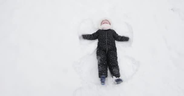Lycklig flicka ligger i snön och gör en ängel — Stockvideo