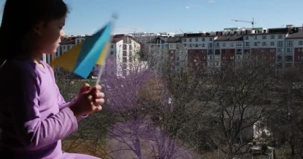 Ein Mädchen mit ukrainischer Flagge sitzt am Fenster, bedeckt mit einer Decke. — Stockvideo