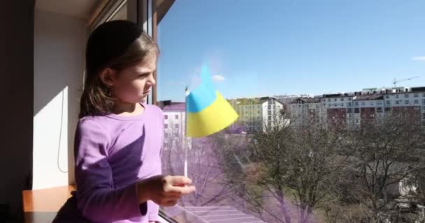 Ein Mädchen mit ukrainischer Flagge sitzt am Fenster, bedeckt mit einer Decke. — Stockvideo