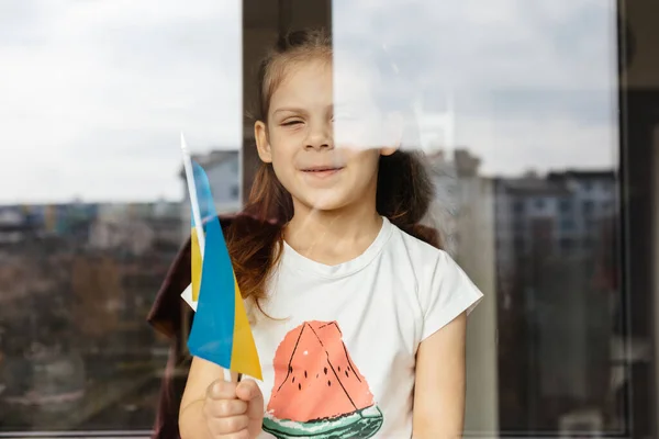 Chica con la bandera de Ucrania en sus manos mira por la ventana — Foto de Stock