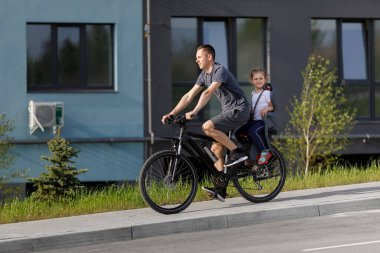 Baba bisiklete biniyor. Küçük kız çocuk koltuğunda.