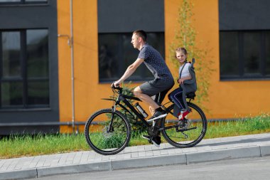 Baba bisiklete biniyor. Küçük kız çocuk koltuğunda.