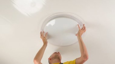A young man, an electrician, is fixing a chandelier, he screws a fixing screw into the base of the lamp in the ceiling. The repair of an apartment.
