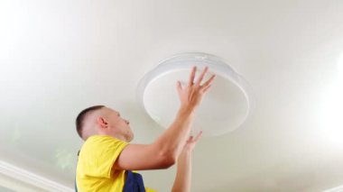 A young man, an electrician, is fixing a chandelier, he screws a fixing screw into the base of the lamp in the ceiling. The repair of an apartment.