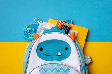 Mavi ve sarı arka planda okul malzemeleriyle dolu bir sırt çantası. Yukarıdan bak. kopyalama alanı. Okul konseptine dönelim..