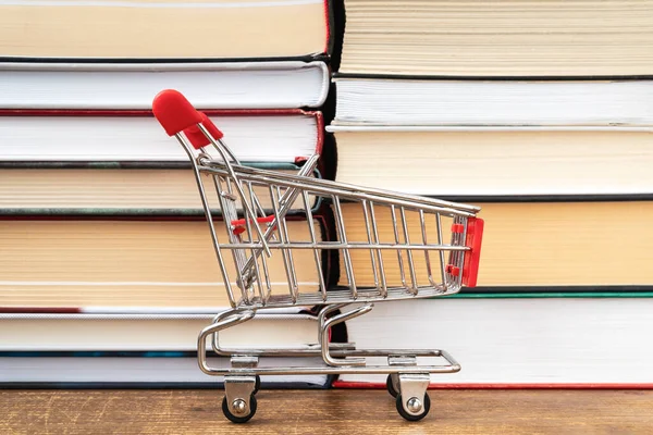 An empty shopping cart stands against the background of books. The concept of buying books in a bookstore or online.
