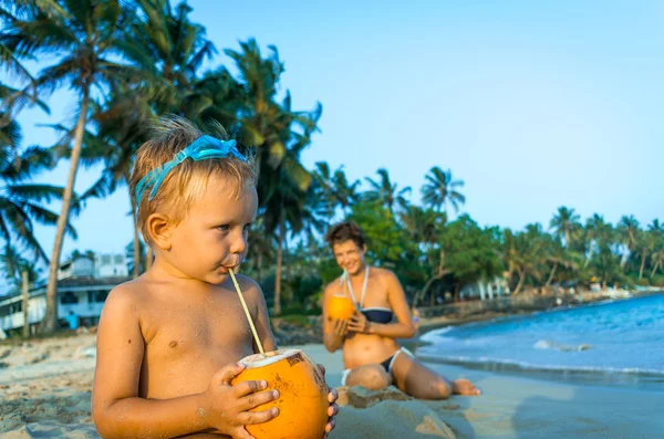 Piccolo ragazzo seduto sulla spiaggia e bere latte di cocco — Foto Stock