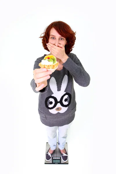 Young woman standing on the scale, holding a cake and closing he — Stock Photo, Image