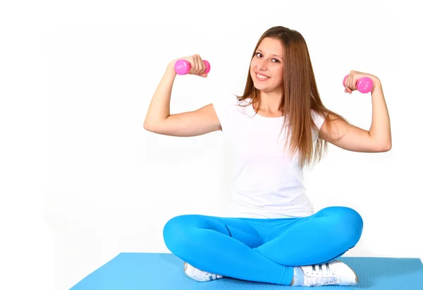 Smiling girl sitting on the mat with the dumbbells isolated on w — Stock Photo, Image