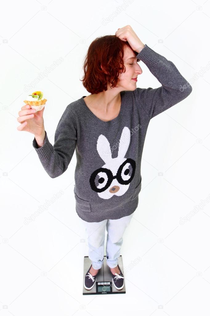 Young woman standing on the scales with a cake and turns away is