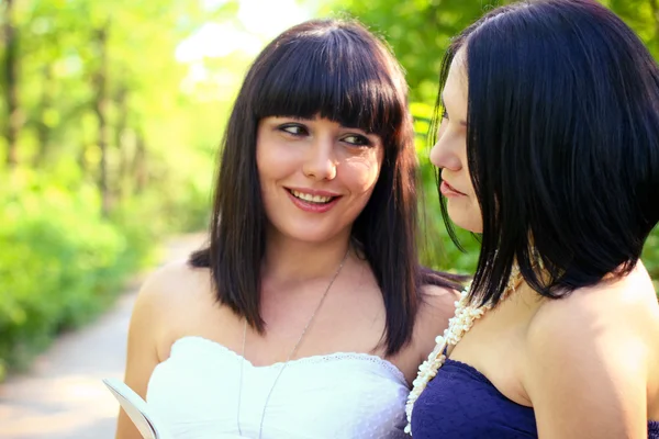 Zwei lächelnde brünette Frauen, die in einem Sommerpark ein Buch lesen — Stockfoto