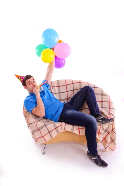 Guy sitting on the couch with a cap and balloons and whistling — Stock Photo, Image