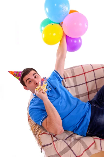 Guy sitting on the couch with a cap and balloons and whistling — Stock Photo, Image