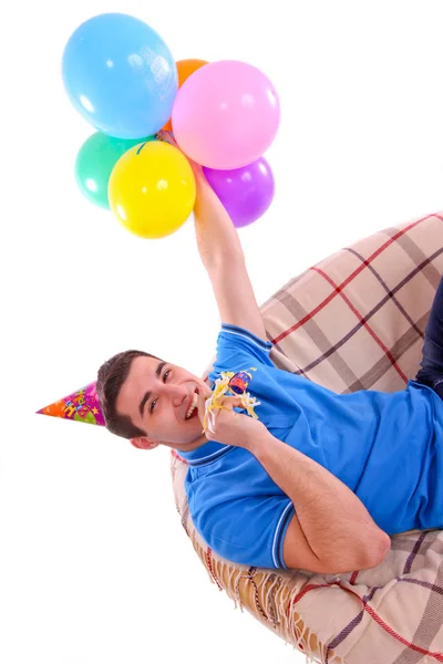 Guy sitting on the couch with a cap and balloons and whistling — Stock Photo, Image