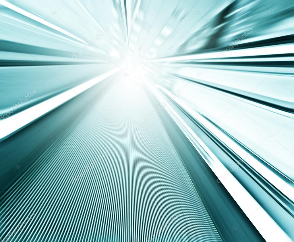 Perspective wide angle view of modern light blue illuminated and spacious high-speed moving commercial escalator with fast blurred trail of handrail in vanishing traffic motion in airport corridor