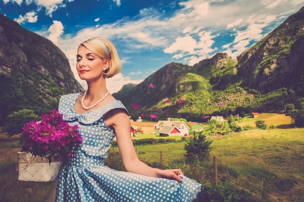 Mulher encantadora em vestido azul com cesta de flores contra vista pequena aldeia — Fotografia de Stock