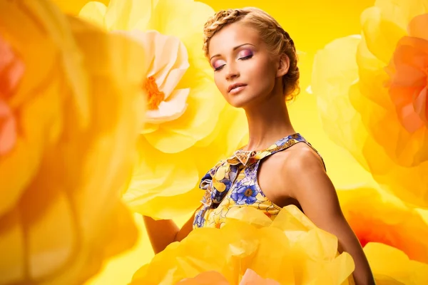 Schöne junge blonde Frau mit geschlossenen Augen in buntem Kleid zwischen großen gelben Blumen — Stockfoto