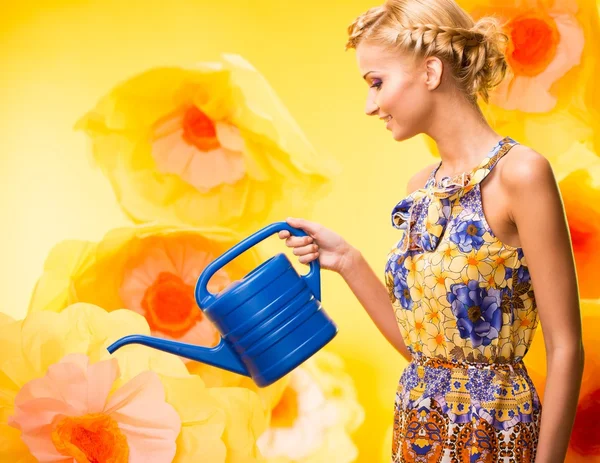Mooie jonge vrolijke blonde vrouw in kleurrijke jurk onder grote gele bloemen met gieter — Stockfoto
