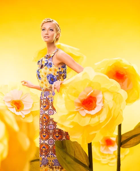 Schöne junge blonde Frau in buntem Kleid zwischen großen gelben Blumen — Stockfoto