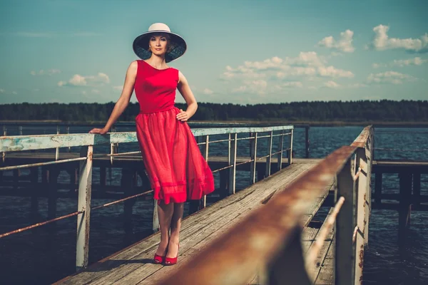 Stylische Frau mit weißem Hut und rotem Kleid steht auf einem alten Holzsteg — Stockfoto