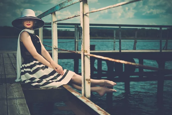 Beautiful woman wearing hat and white scarf sitting on old wooden pier — Stock Photo, Image