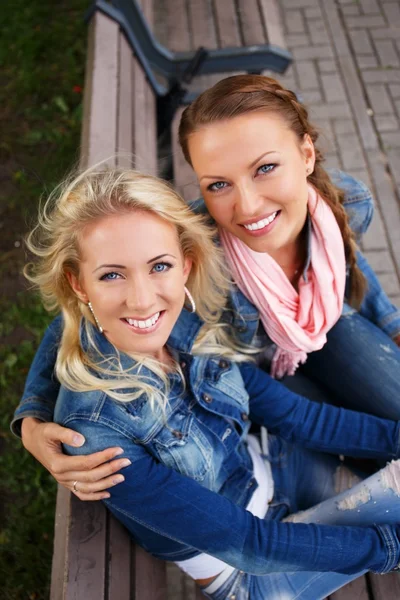 Zwei schöne lächelnde junge Mädchen in Jeansjacken sitzen auf einer Bank in einem Park — Stockfoto