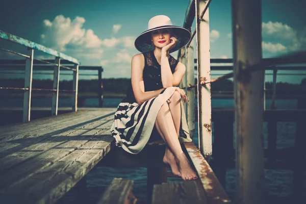 Beautiful woman wearing hat and white scarf sitting on old wooden pier — Stock Photo, Image