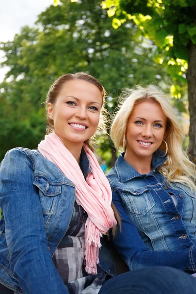 Två vackra leende ung tjej i jeans jackor satt på en bänk i en park — Stockfoto