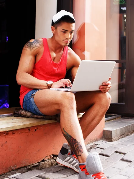 Uomo in pantaloncini e berretto seduto con un computer portatile — Foto Stock