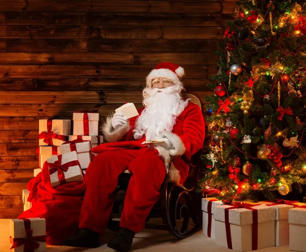 Άγιος Βασίλης κάθεται στο ρηλάξ στο ξύλινο σπίτι εσωτερικό με γράμματα στα χέρια — Φωτογραφία Αρχείου