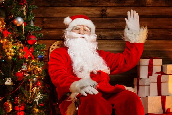 Santa claus sedí na houpací křeslo v dřevěných domů interiéru s dárkové krabice kolem něj — Stock fotografie