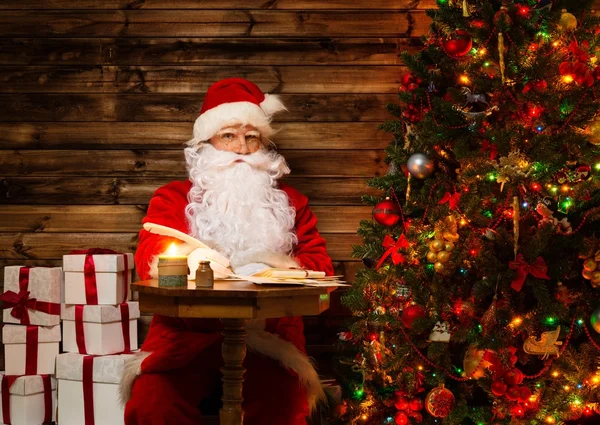 Santa Claus in wooden home interior sitting behind table and writing letters with quill pen — Stock Photo, Image