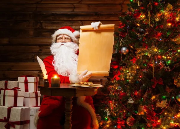 Papai Noel em casa de madeira interior segurando rolagem lista de desejos em branco — Fotografia de Stock