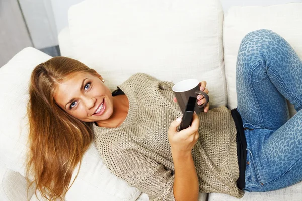 Giovane bella donna su un divano con telecomando e tazza — Foto Stock