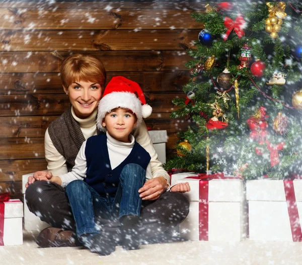 Madre y su pequeño niño en Santa sombrero con caja de regalo bajo el árbol de Navidad en el interior de la casa de madera —  Fotos de Stock