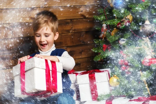 Kleiner Junge öffnet Geschenkbox unter Weihnachtsbaum im Holzhaus-Interieur — Stockfoto