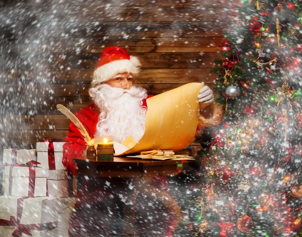 Santa Claus in wooden home interior reading wish list scroll — Stock Photo, Image