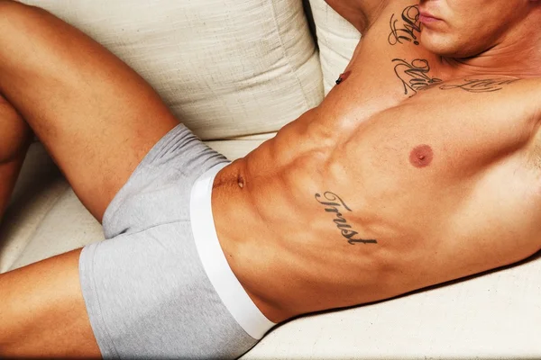 Hombre con hermoso torso musculoso tatuado en ropa interior tumbado en el sofá —  Fotos de Stock