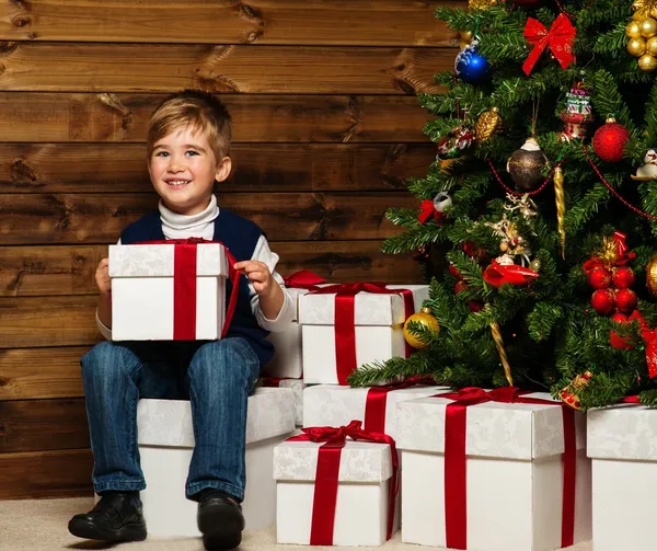 Kleiner Junge mit Geschenkbox unter dem Weihnachtsbaum im Holzhaus-Interieur — Stockfoto