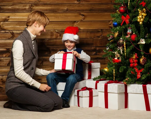 Madre y su pequeño niño en la caja de regalo de apertura de Santa sombrero bajo el árbol de Navidad en el interior de la casa de madera —  Fotos de Stock