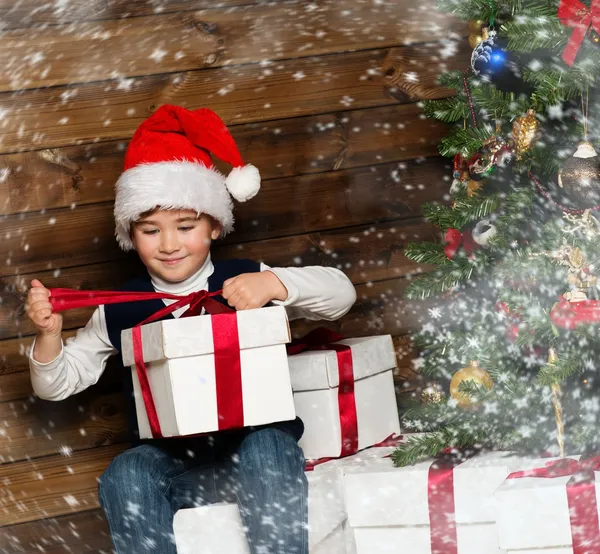 Kleiner Junge mit Weihnachtsmütze öffnet Geschenkbox unter Weihnachtsbaum im Holzhaus-Interieur — Stockfoto