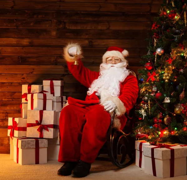 Weihnachtsmann auf Schaukelstuhl im hölzernen Hausinneren sitzend mit Glöckchen in der Hand — Stockfoto