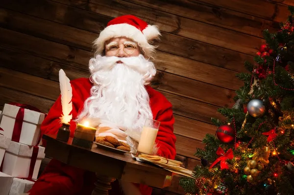 Santa claus in houten huis interieur vergadering achter tafel met melk en havermout cookies — Stockfoto