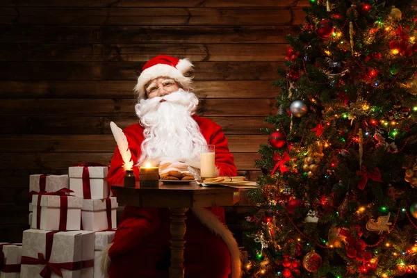 Weihnachtsmann im hölzernen Heim-Interieur sitzt hinter einem Tisch mit Milch und Haferflocken-Keksen — Stockfoto