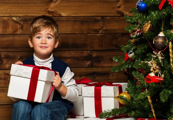 Kleiner Junge mit Geschenkbox unter dem Weihnachtsbaum im Holzhaus-Interieur — Stockfoto