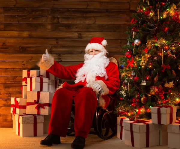 Papai Noel sentado na cadeira de balanço com polegar para cima no interior de casa de madeira com caixas de presente em torno dele — Fotografia de Stock