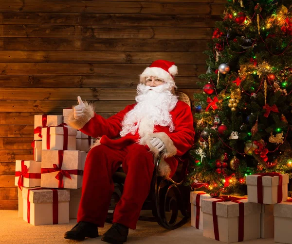 Santa Claus sentado en mecedora con el pulgar hacia arriba en el interior de la casa de madera con cajas de regalo a su alrededor —  Fotos de Stock
